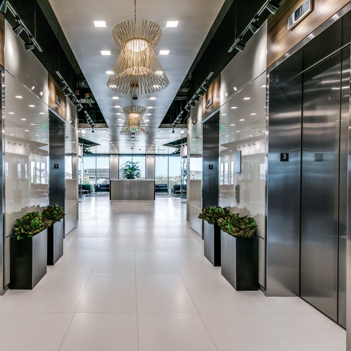 A modern office lobby with two rows of elevators, sleek lighting fixtures above, and potted plants along the sides.