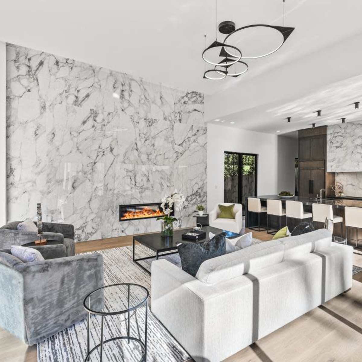 A modern living room featuring a marble accent wall, a sleek fireplace, and an open kitchen area with minimalist furniture and neutral decor.