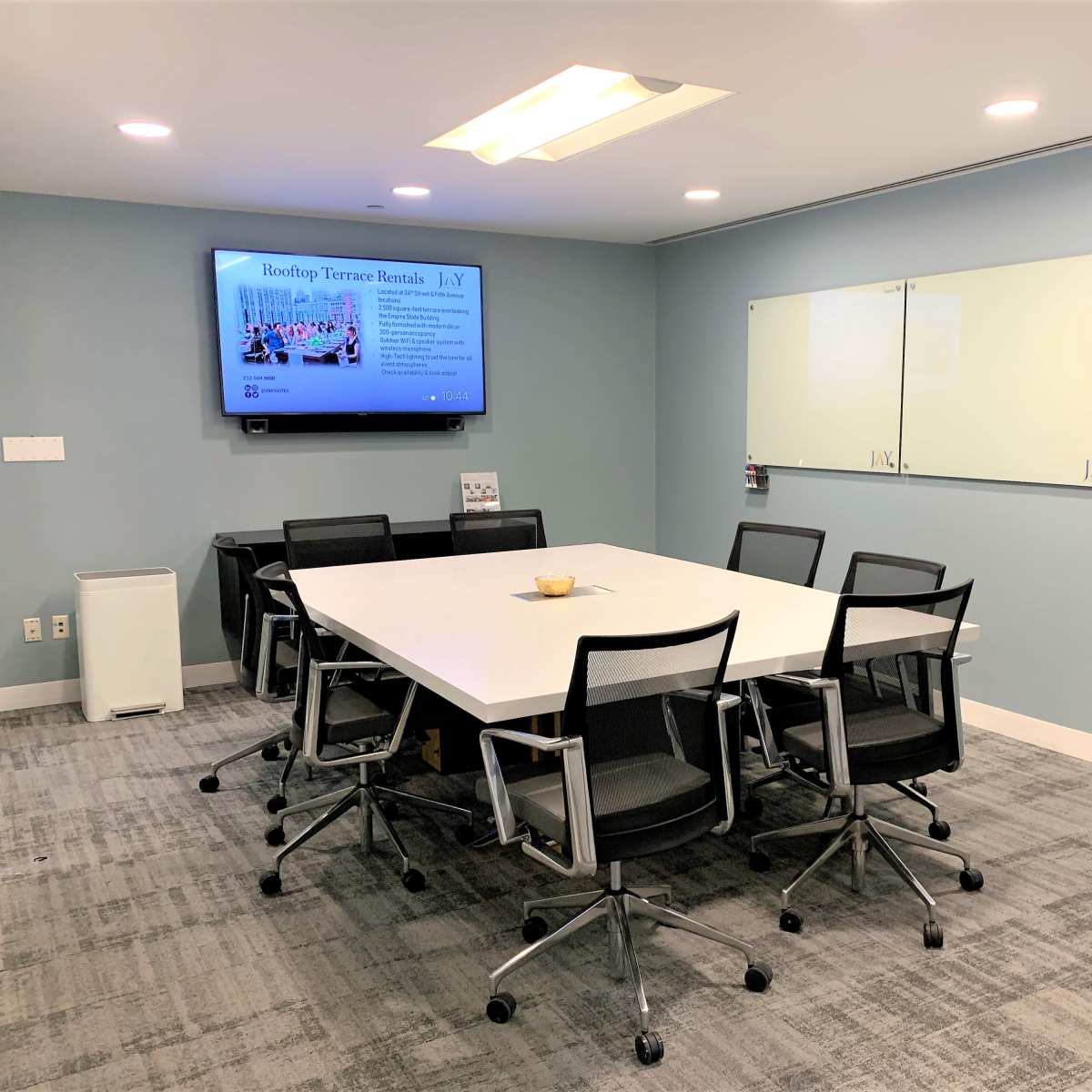 A modern conference room features a large white table surrounded by black chairs, with a wall-mounted screen displaying a presentation and two whiteboards.