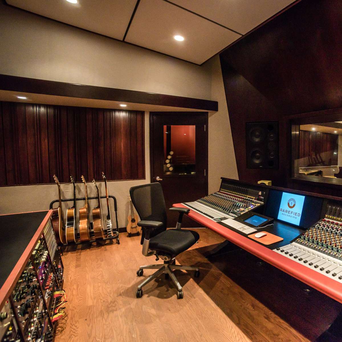 A recording studio features a large mixing console, surrounded by acoustic panels and guitars in a stand.