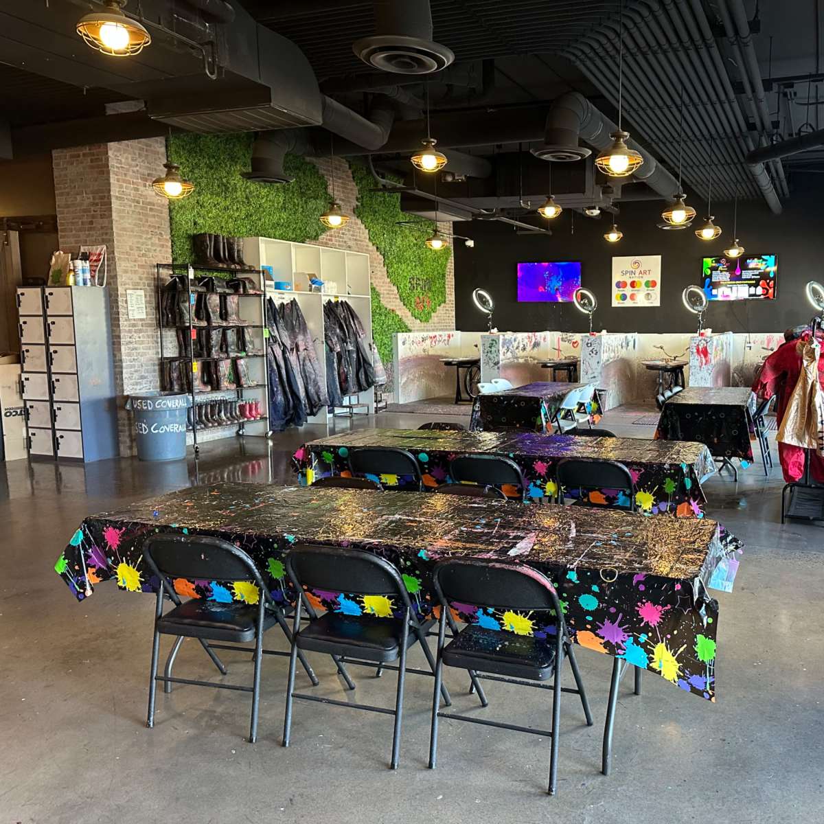 A spacious art studio featuring multiple tables with colorful tablecloths, several chairs, and a backdrop of artwork along the walls.