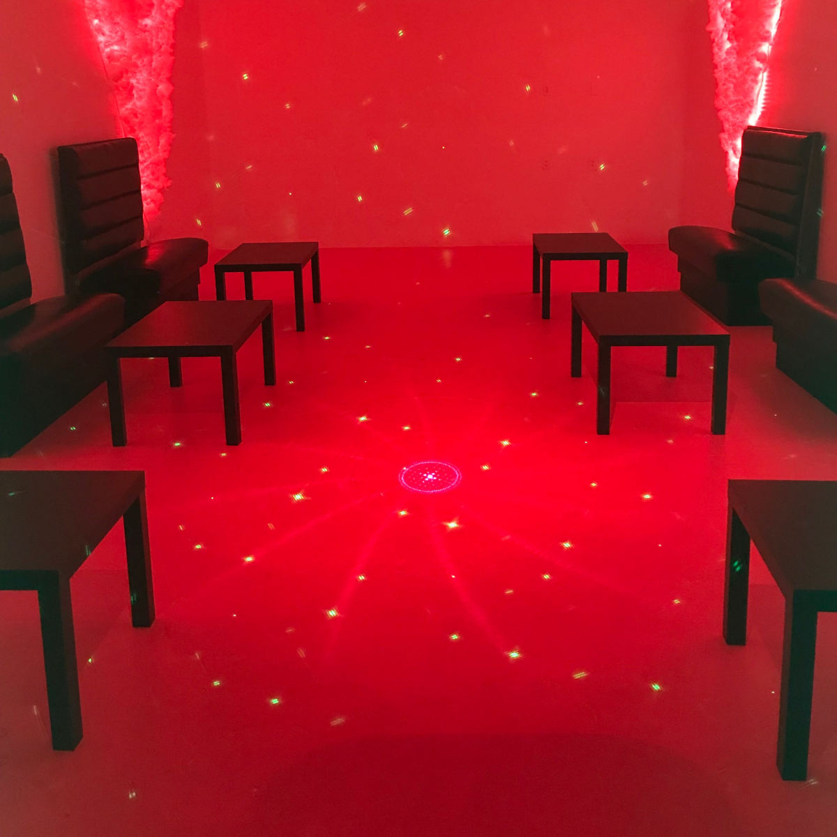 A room with red lighting, featuring black seating arrangements and small tables arranged in a circular formation on a carpeted floor.