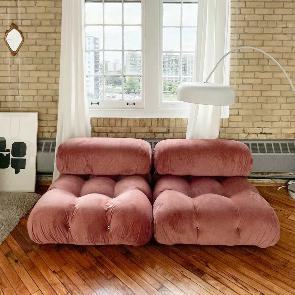 A pair of pink, plush sofas placed near large windows, with sheer white curtains and a standing lamp nearby.
