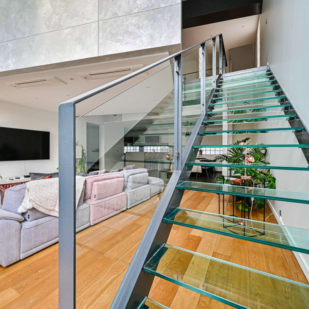 A modern interior features a glass staircase leading to an upper level, with a spacious living area that includes a large sofa and a television.