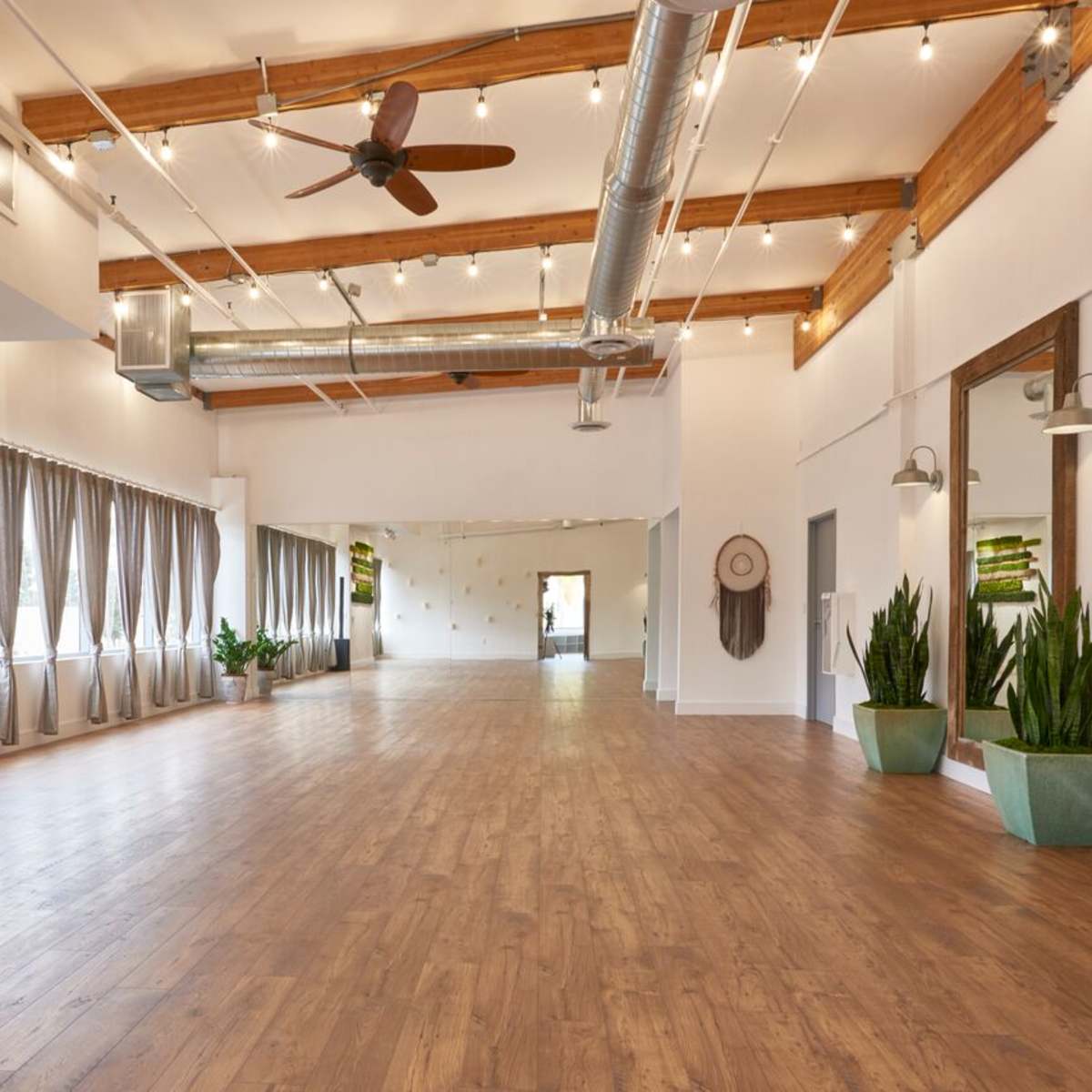 A spacious, well-lit interior room with wooden flooring, decorative plants, and large mirrors on the walls.