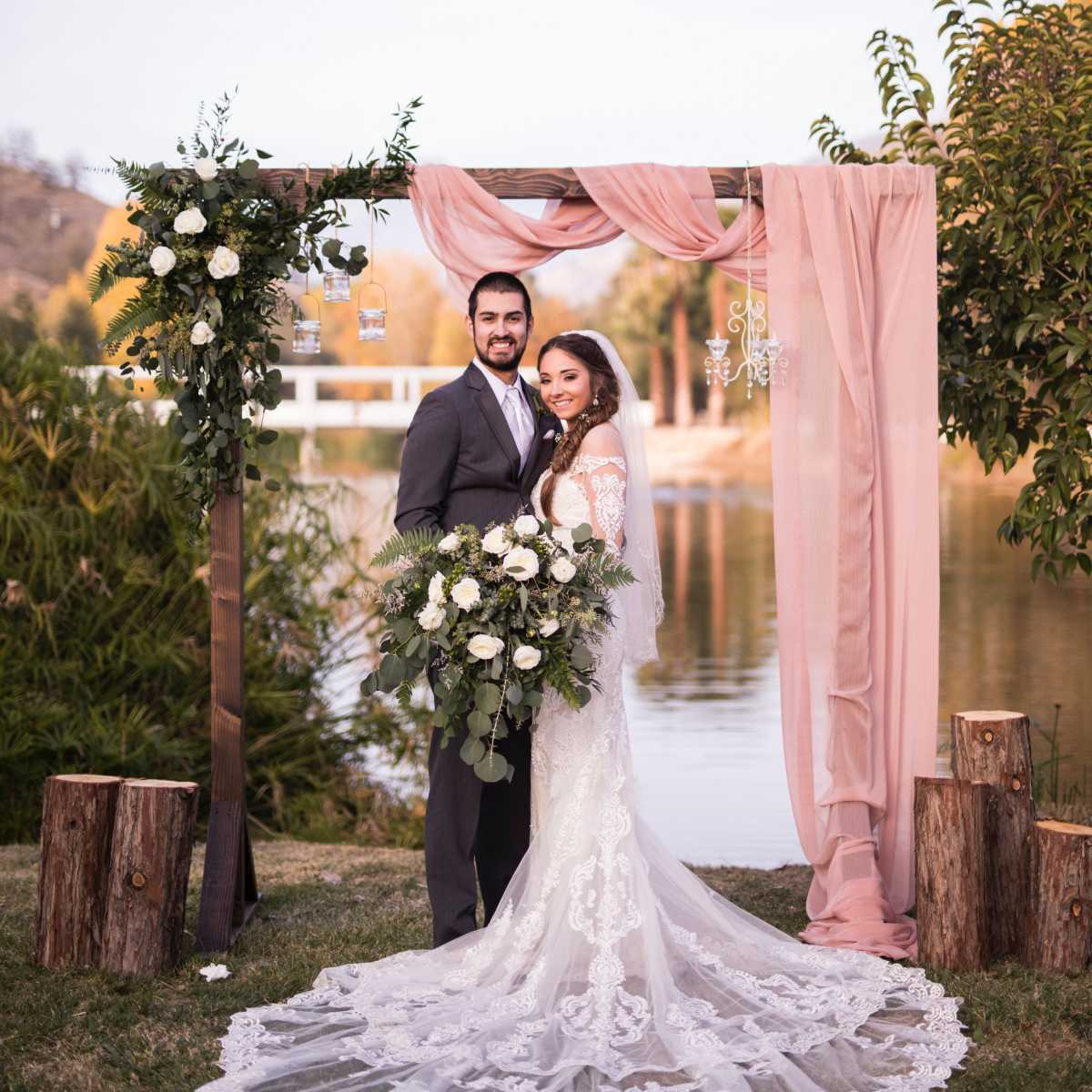 A bride and groom stand together under a floral arch beside a tranquil body of water.
