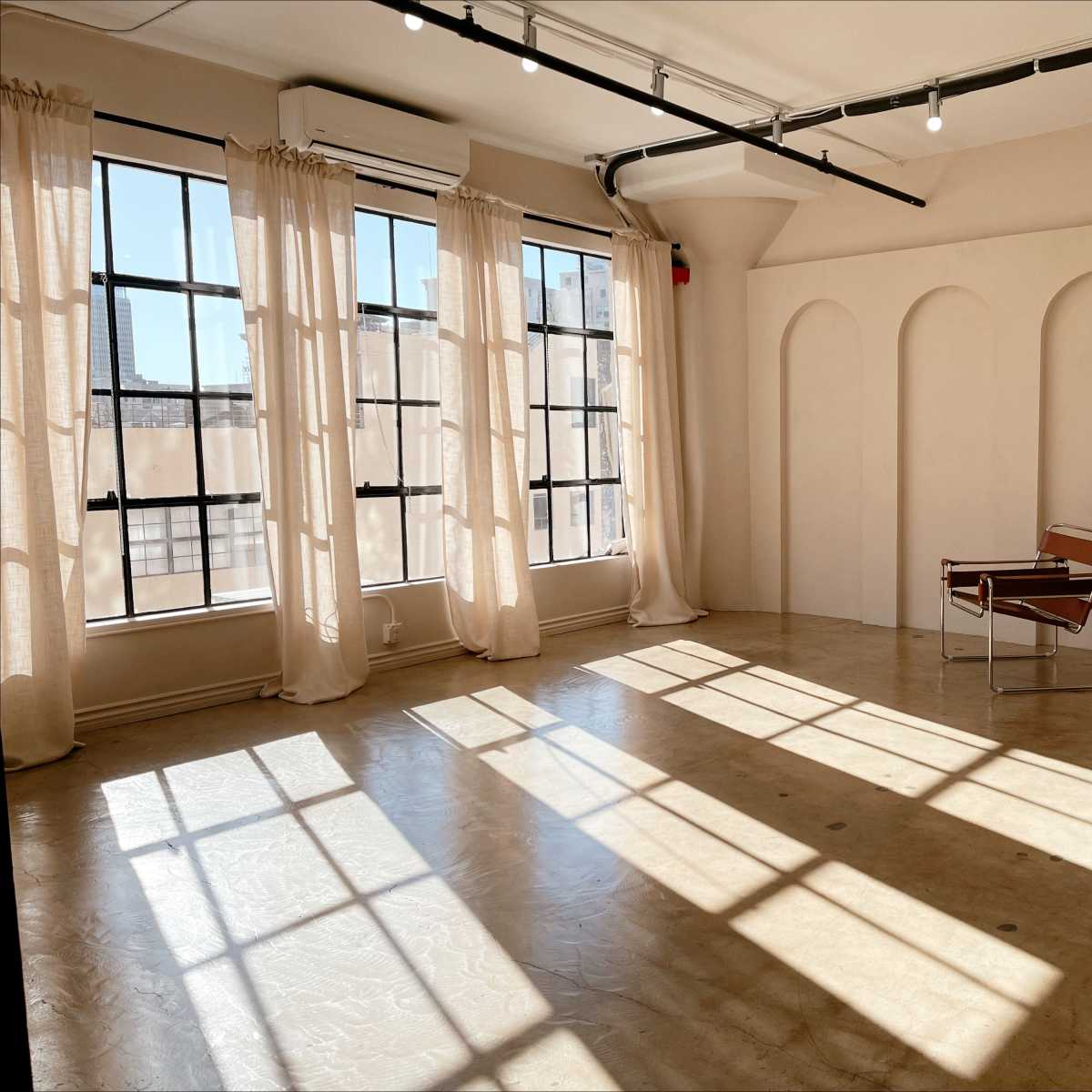 A well-lit, minimalist interior with large windows, sheer curtains, and a modern chair positioned on a polished concrete floor.