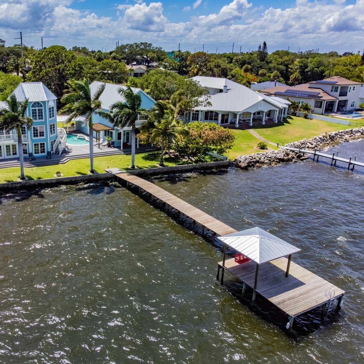 A waterfront property features two distinct homes, a wooden dock extending into the water, and palm trees lining the shoreline.