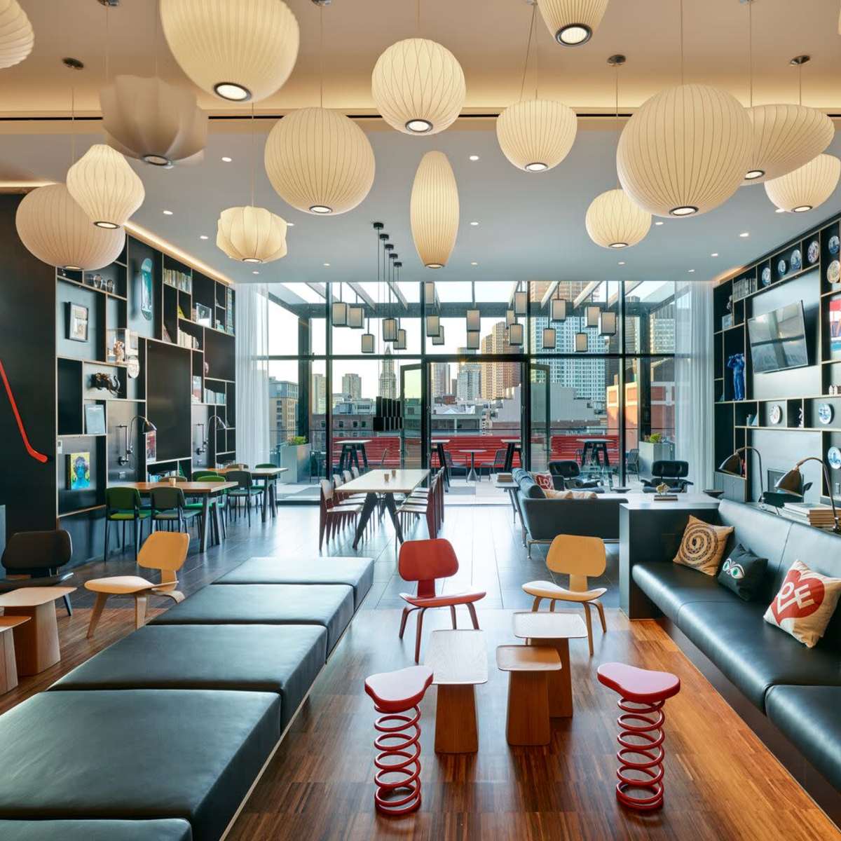 A modern lounge area with black seating, colorful stools, and decorative pendant lights, featuring large windows that provide a view of the city.
