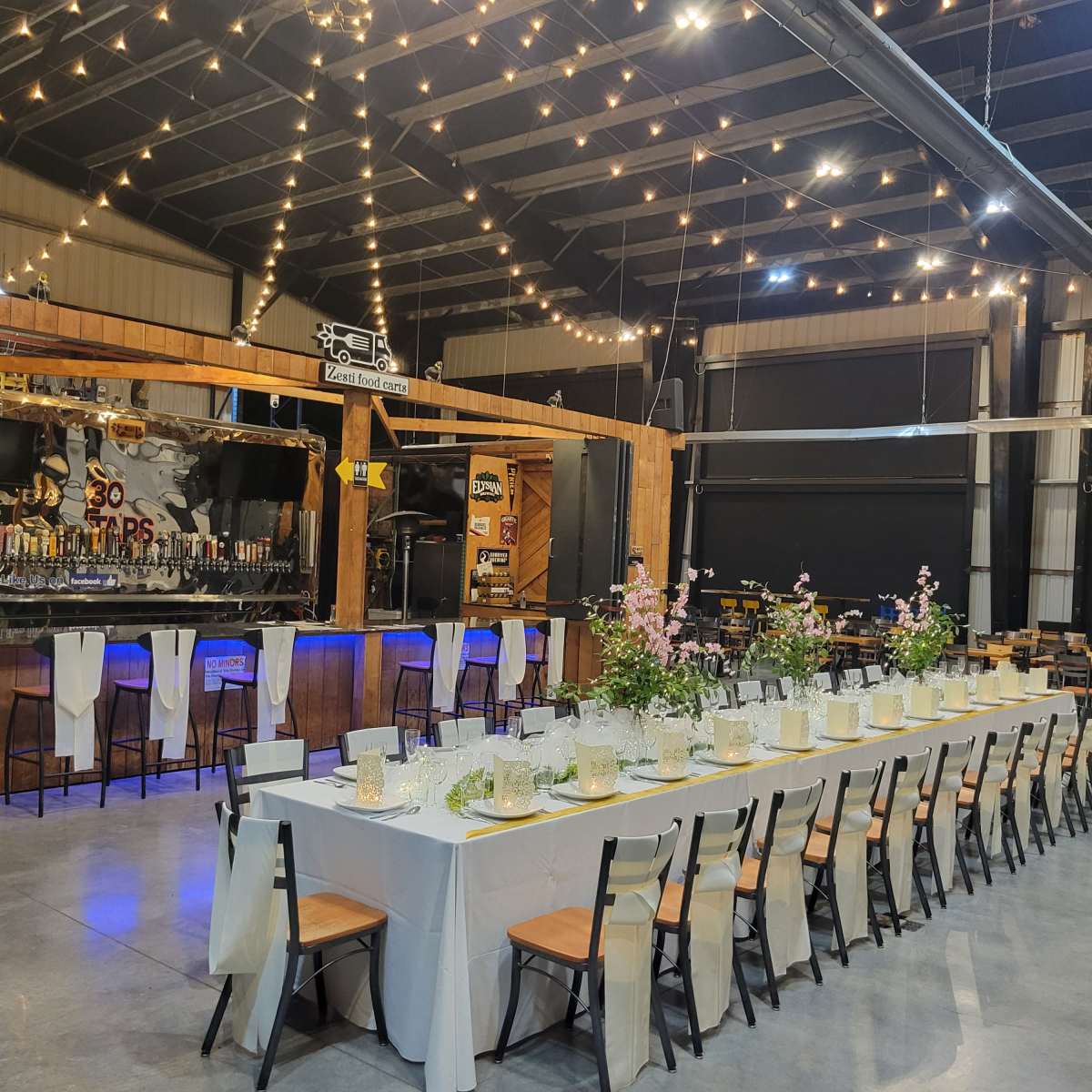 A long dining table adorned with flowers and candles is set up in a spacious venue with string lights overhead and a bar in the background.