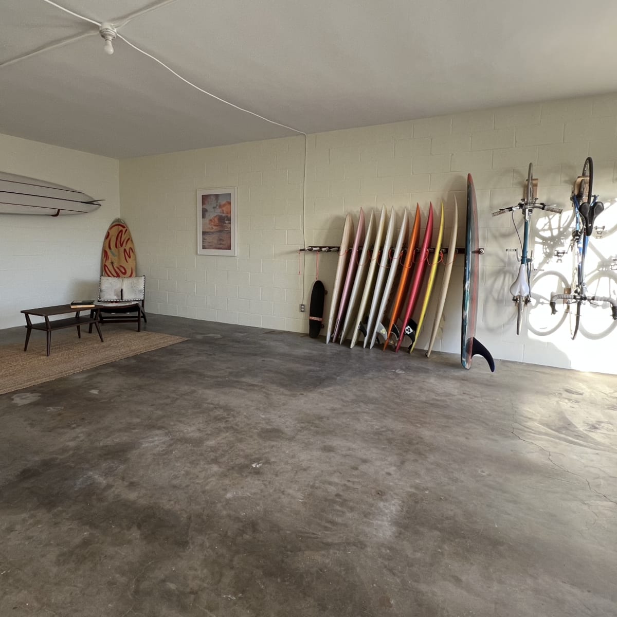 A spacious garage with surfboards and bicycles neatly arranged on the walls and a seating area consisting of a chair and small table.