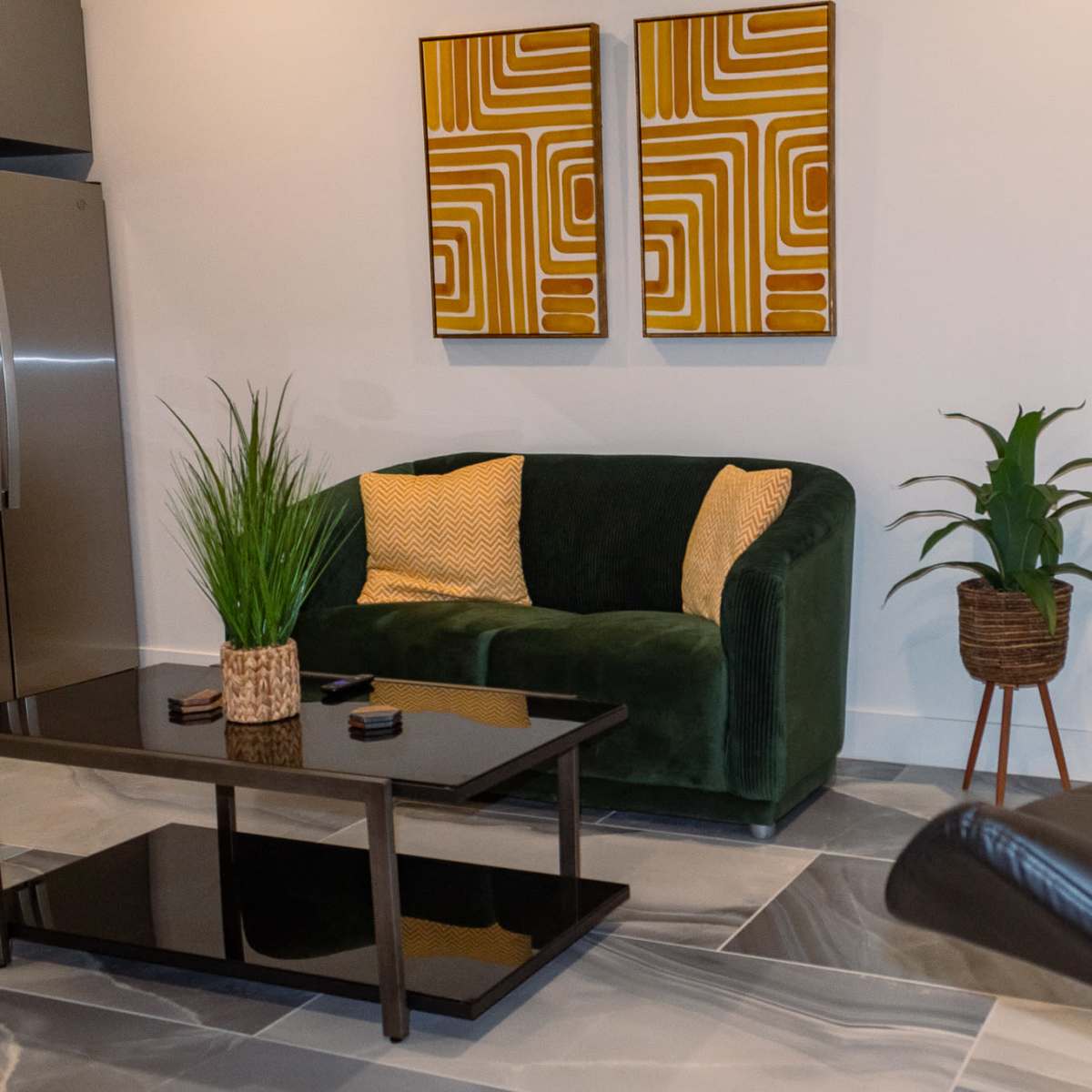 A modern living area featuring a green sofa, two abstract art pieces on the wall, a glass-topped coffee table, and a small round dining table with black chairs.