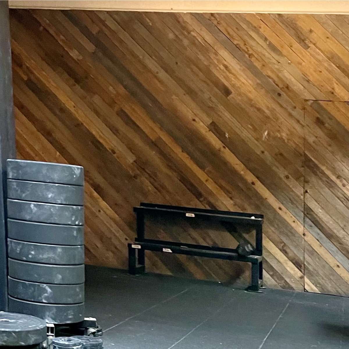A gym setting with a stacked weight plate on the left and a black bench positioned against a wooden diagonal-paneled wall.