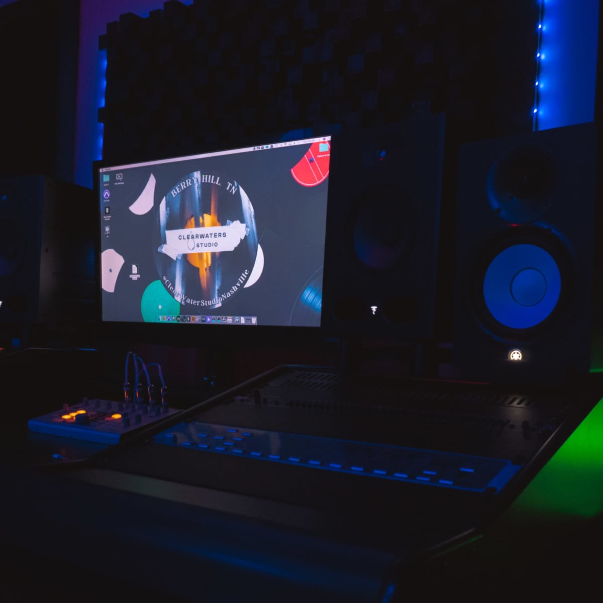 A music studio with a computer display showing a digital audio workstation, flanked by speakers and a mixing console, illuminated by ambient lighting.
