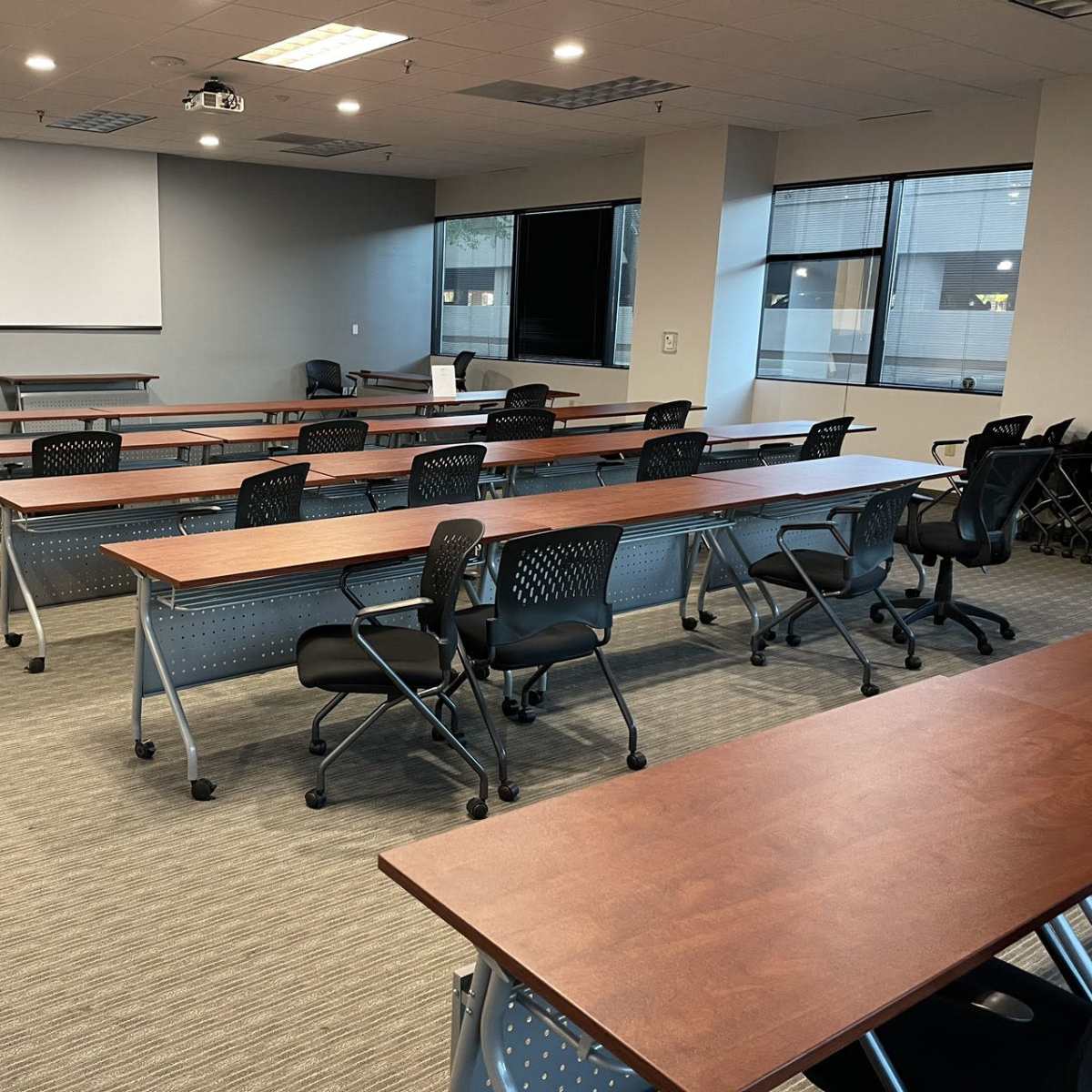 A classroom or meeting room setup with multiple rows of desks and chairs arranged for participants, along with a projector and whiteboard at the front.