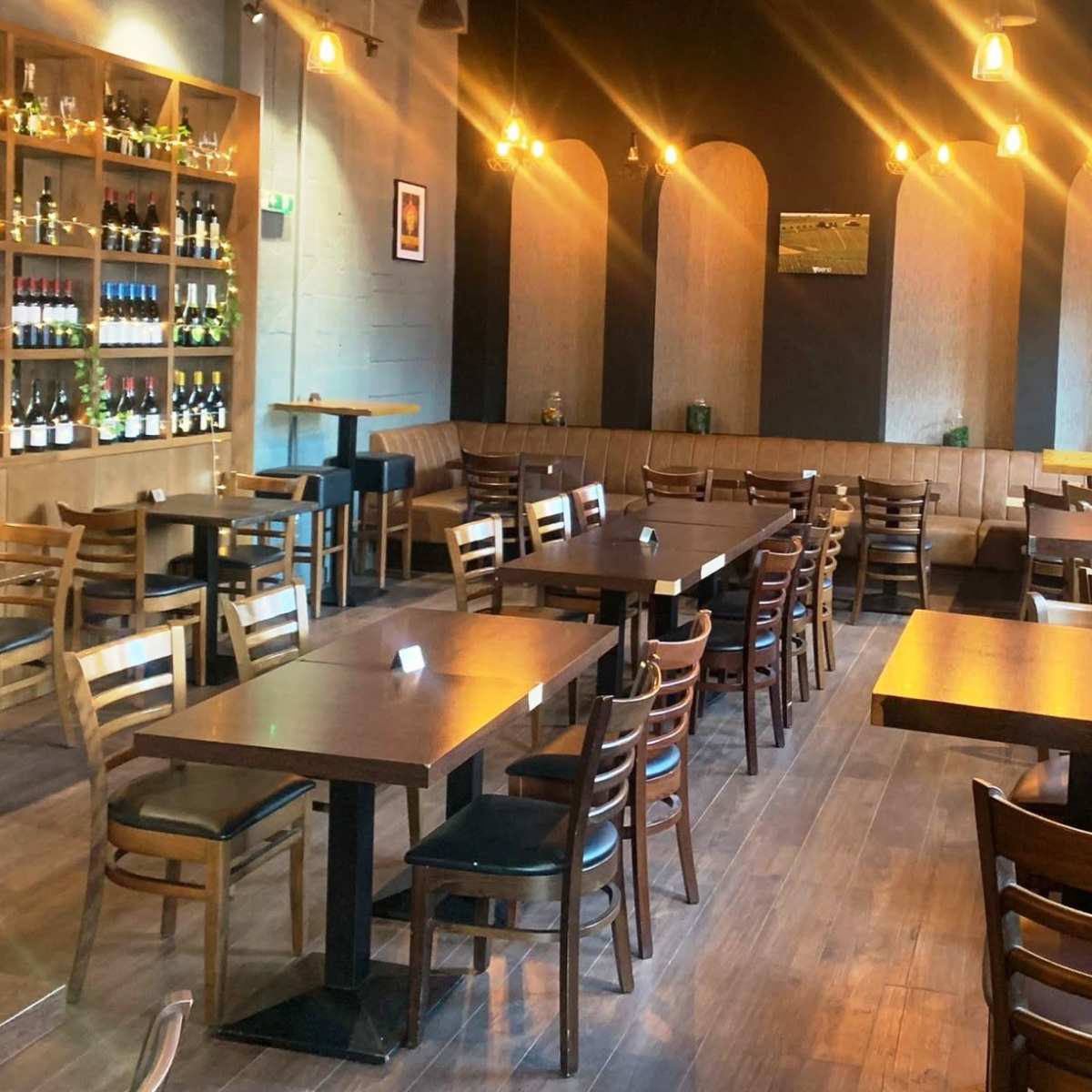 A spacious restaurant interior with wooden tables and chairs, shelves displaying bottles, and warm lighting.