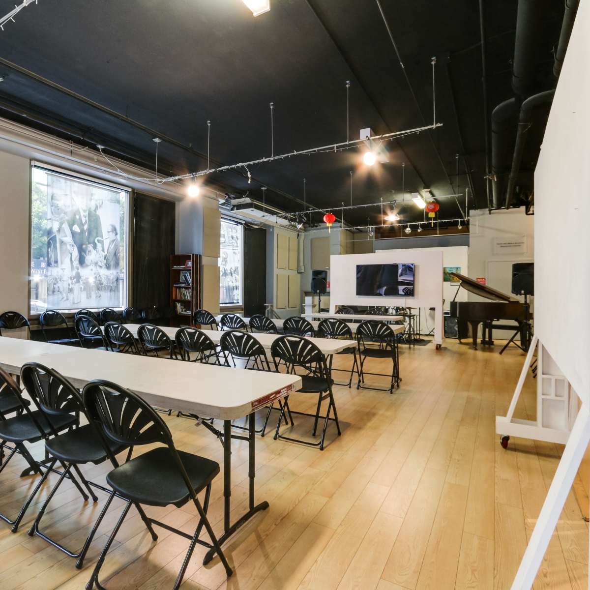A large, open room features rows of black folding chairs facing a central white screen, with windows showcasing the view of a city street.