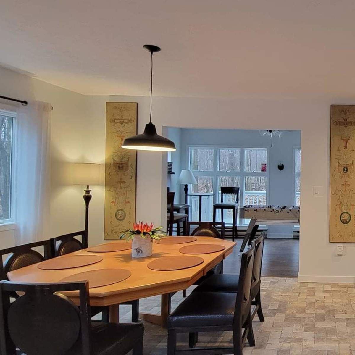 A modern kitchen and dining area with a round wooden table, chairs, and a long counter with stools, all set in a well-lit space with large windows facing a wooded area.