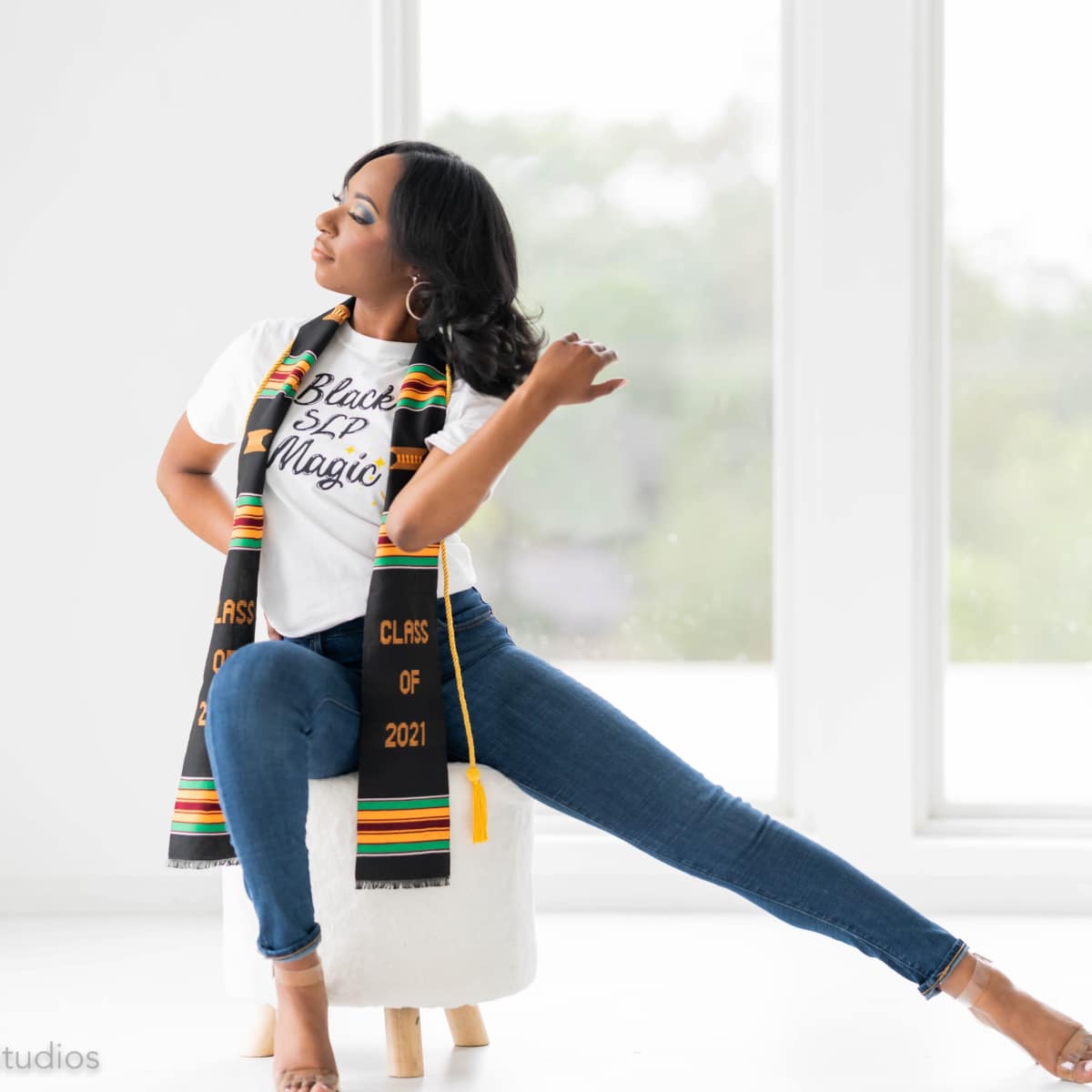 A young woman sits on a stool, wearing a "Black SIP Magic" t-shirt and a graduation scarf, while extending one leg out beside her.
