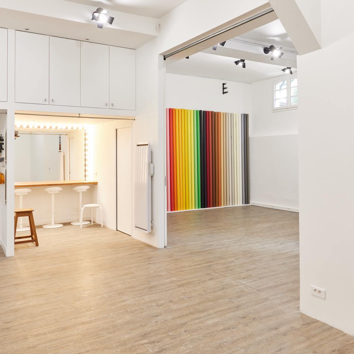 A spacious studio with light wooden flooring, featuring a bar area with two stools and a wall of colorful vertical stripes.