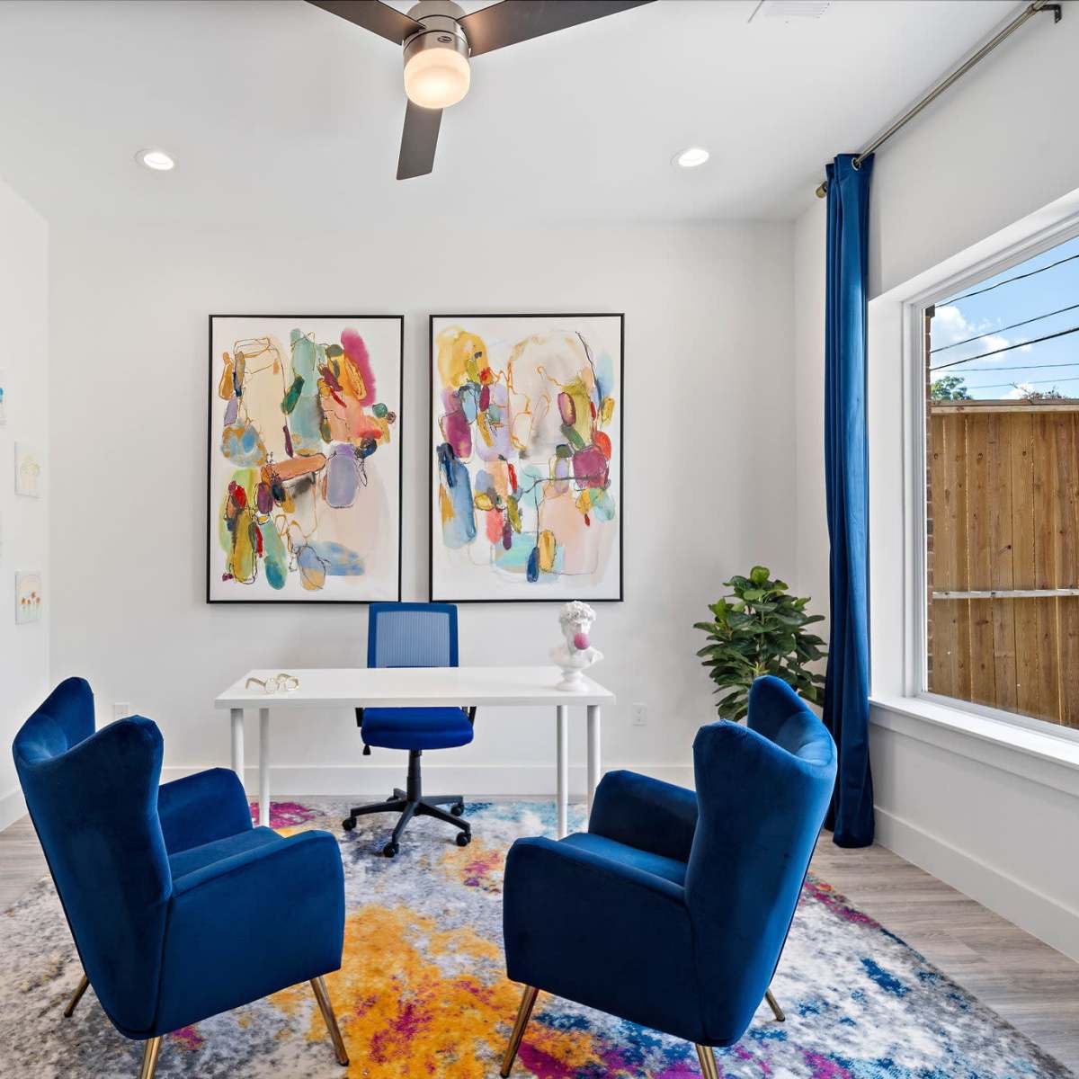 A bright home office features two blue chairs, a white desk, colorful wall art, and large windows with a view of a wooden fence.