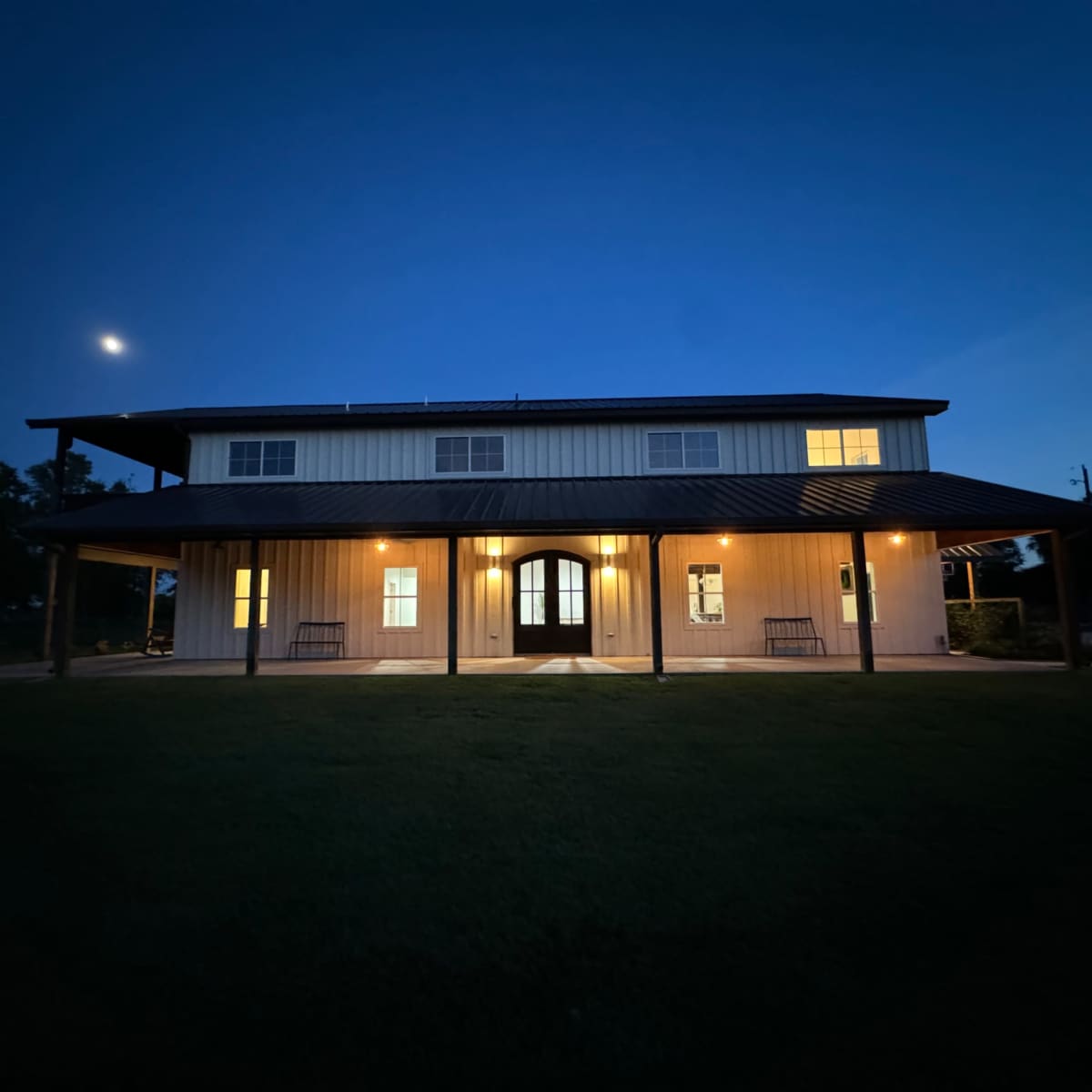 A two-story house with a metal roof and illuminated windows, set in a grassy area at dusk under a clear sky with the moon visible.
