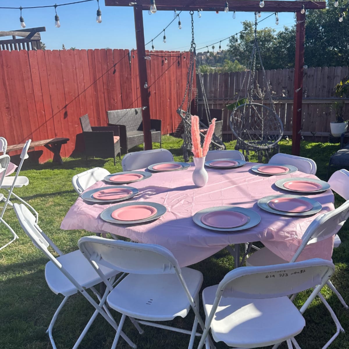 A round table is set with pink plates and a white vase on a grassy area, surrounded by chairs and outdoor furniture beneath string lights.