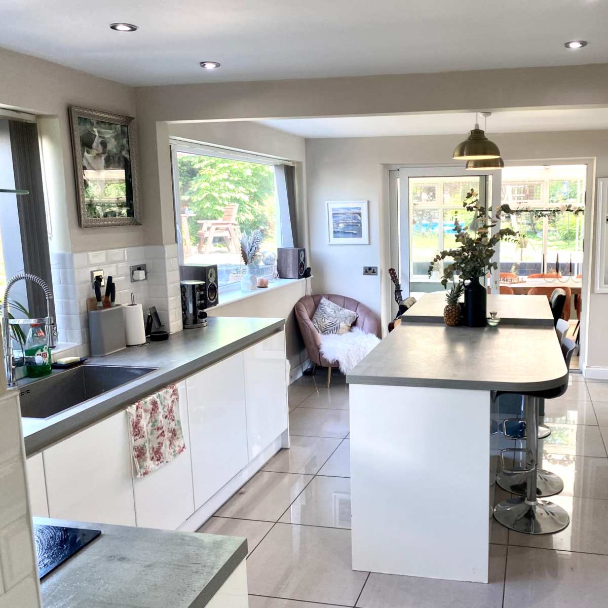 A modern kitchen features sleek white cabinetry, a large island with a gray countertop, and a view of a sunny outdoor seating area through the windows.