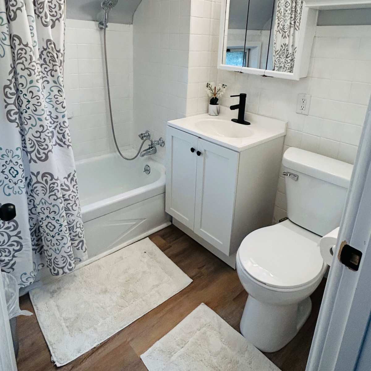 A compact bathroom featuring a bathtub with a shower, a white vanity with a sink, a toilet, and decorative elements on the walls.