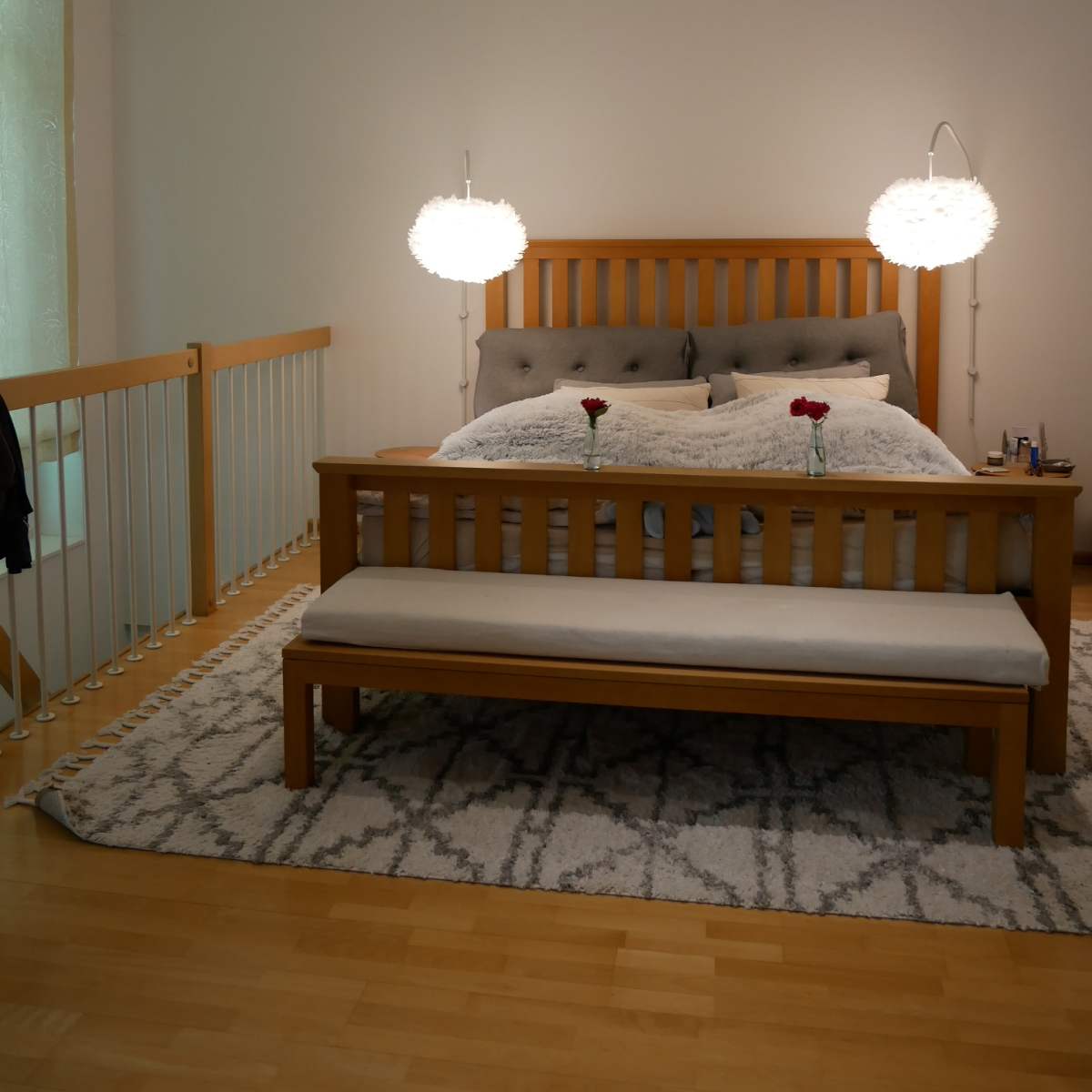 A wooden bed with decorative pillows and roses sits in a softly lit bedroom, accompanied by a bench and two hanging lights.