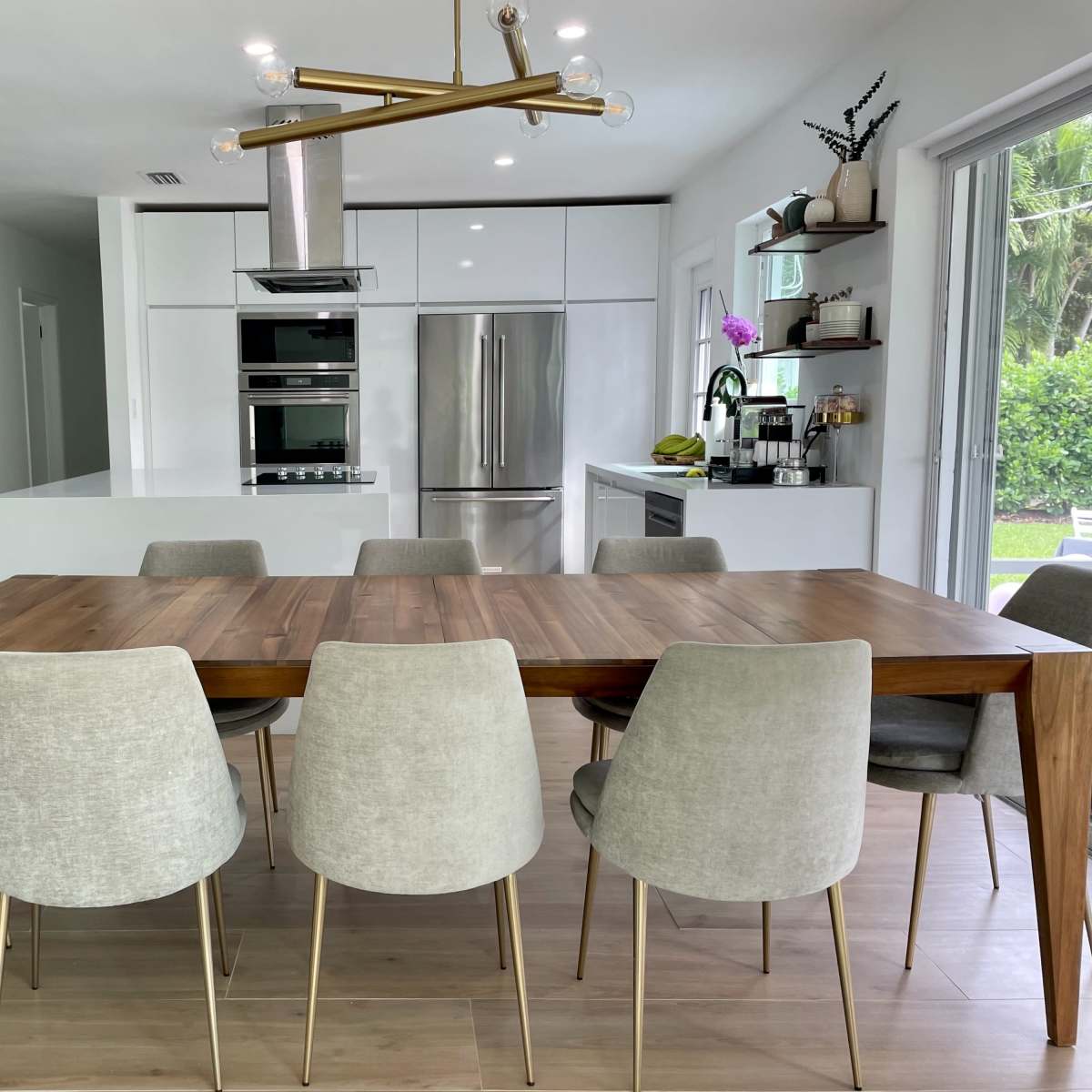 A modern kitchen and dining area featuring a wooden dining table surrounded by gray upholstered chairs, with sleek cabinetry and stainless steel appliances in the background.