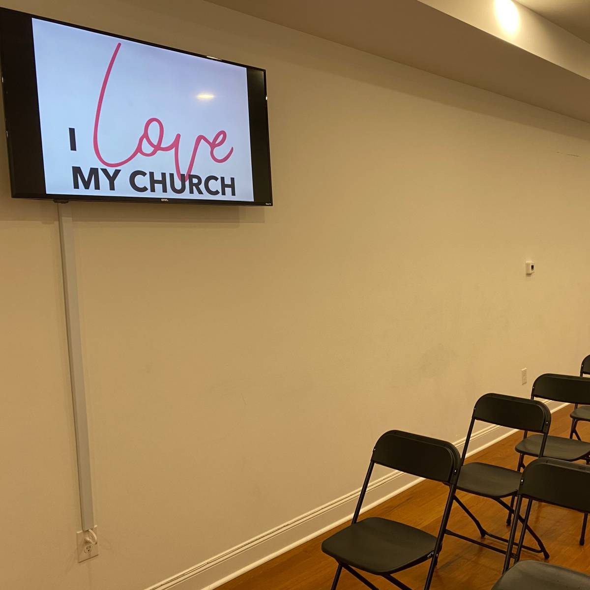 A television screen displaying "I love MY CHURCH" is mounted on a wall, with rows of black folding chairs set up in front of it.