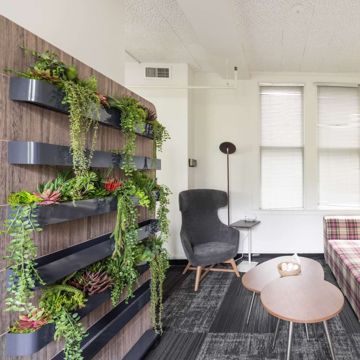 A small lounge area features a plaid sofa, wooden side tables, a modern chair, and a wall adorned with vertical planters.