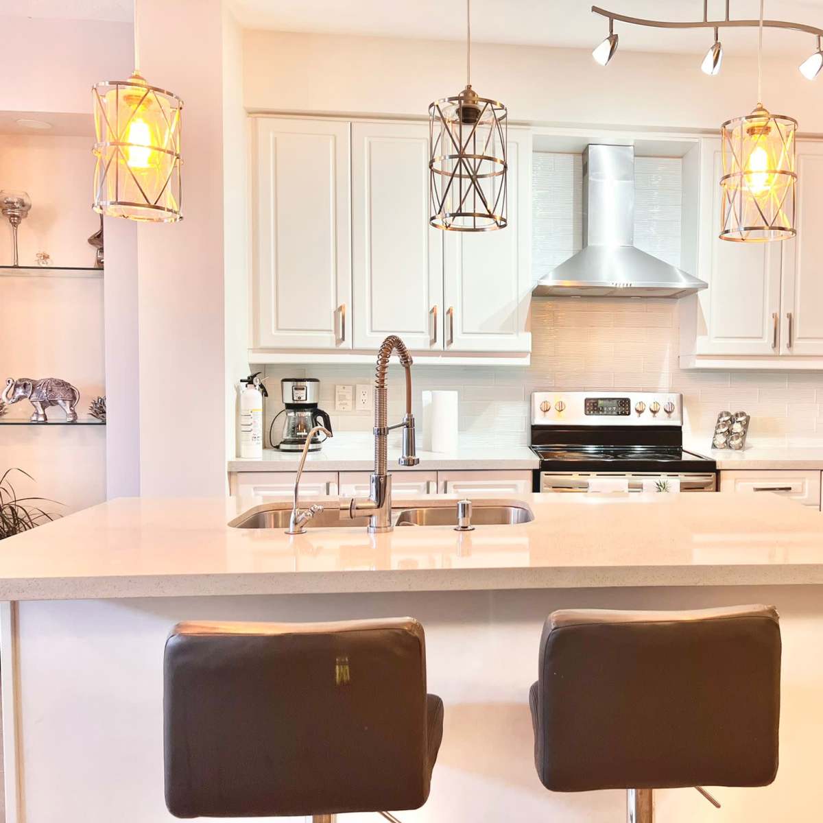 A modern kitchen featuring a central island with two black bar stools, overhead pendant lighting, and sleek white cabinetry.