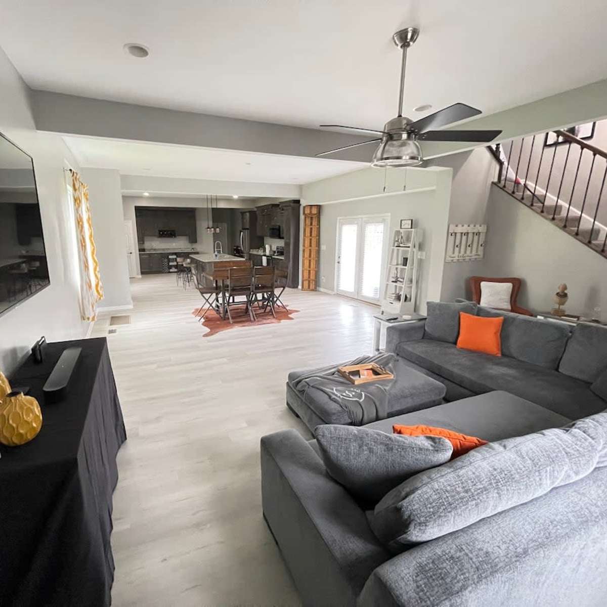 A spacious living area with a gray sectional sofa, a flat-screen TV mounted on the wall, and a dining area visible in the background.
