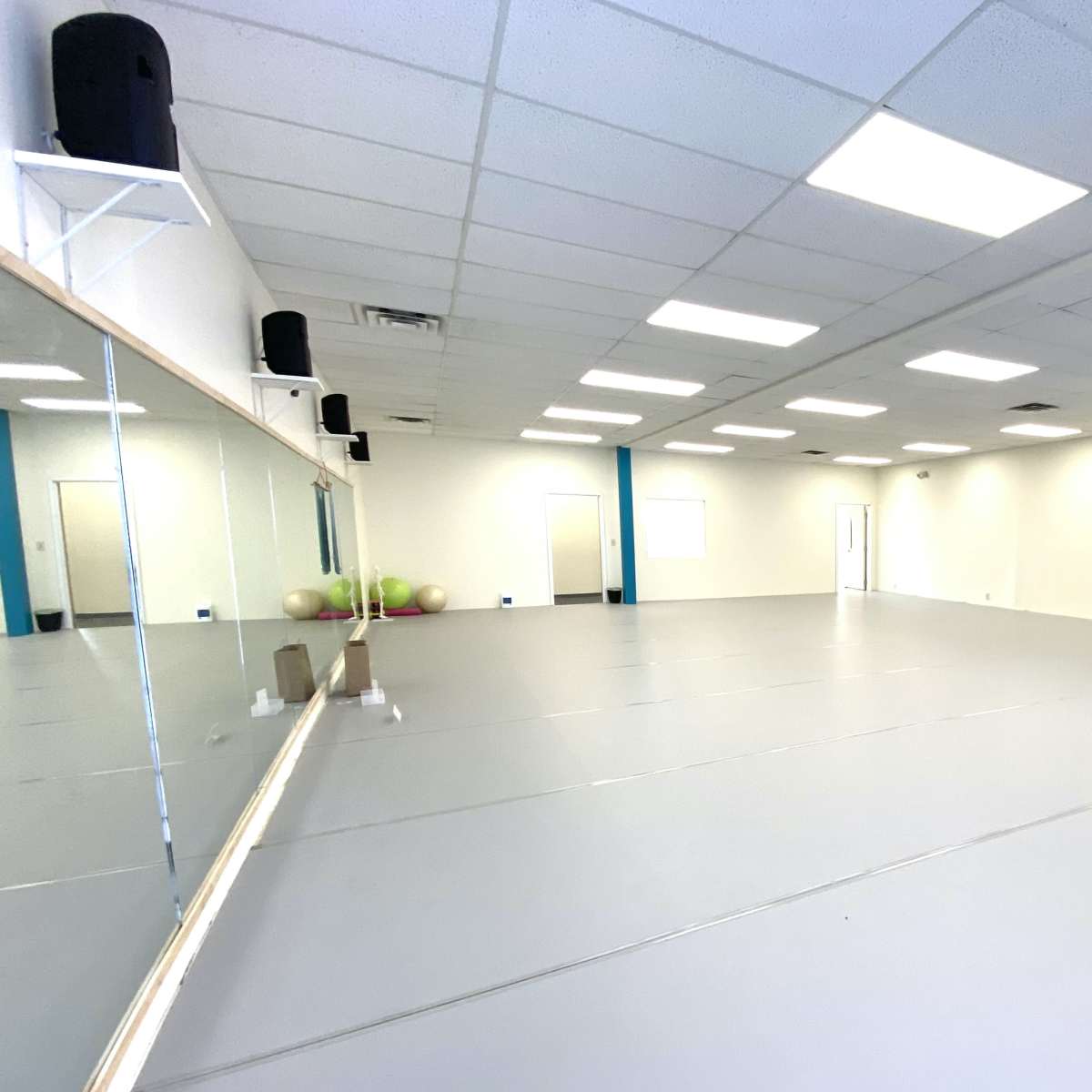 A spacious, empty room with a gray floor, illuminated by overhead lights, featuring a large mirror on one wall and a door on the opposite side.