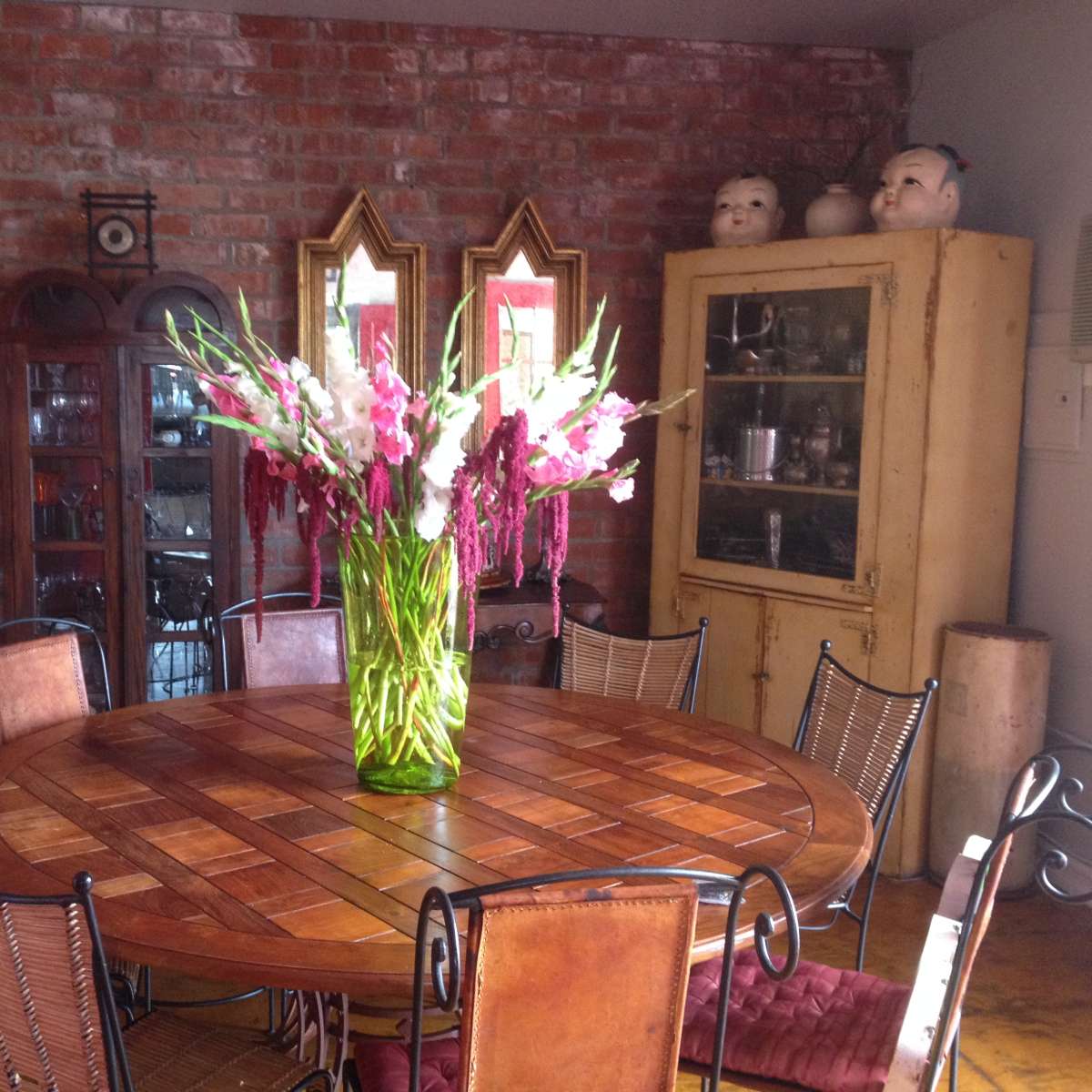 A round wooden dining table is surrounded by wrought iron chairs, topped with a large vase of flowers in a brick-walled room featuring a cabinet and decorative mirrors.