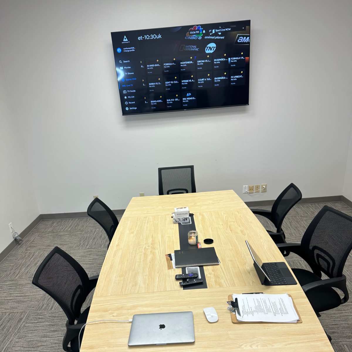A conference room with a wooden table surrounded by six black chairs, featuring a mounted screen displaying a schedule and a laptop on the table.