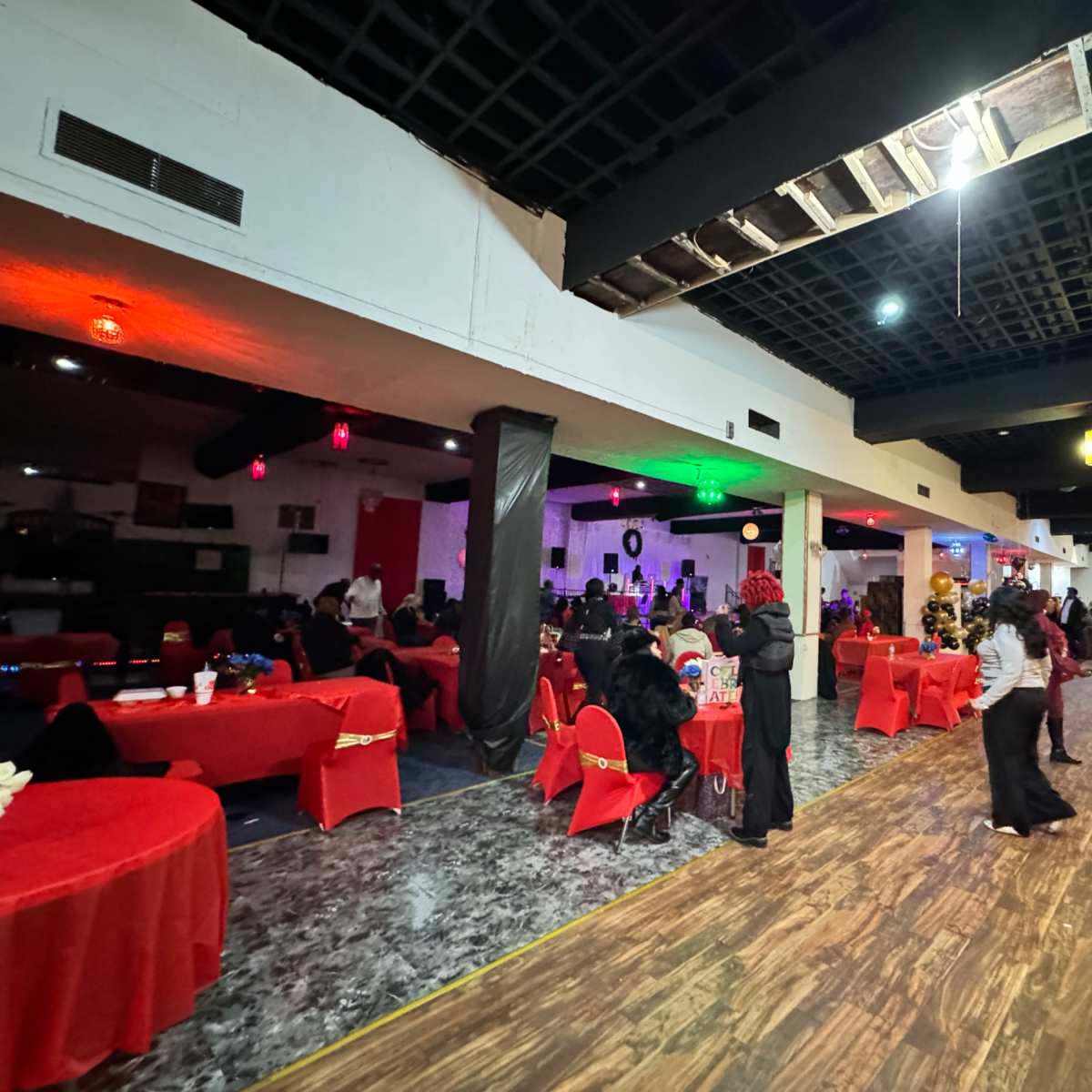 A spacious event hall is filled with tables covered in red tablecloths and attendees in formal attire, while a stage at the back is set up for a performance.