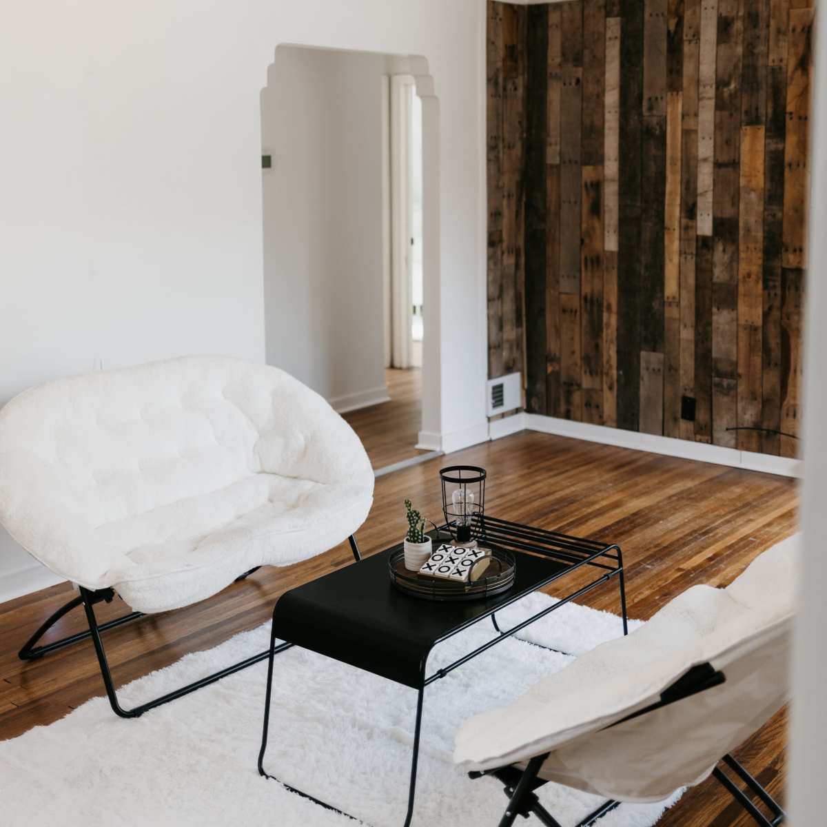 A living room with two white chairs, a black table, and wood-paneled walls, illuminated by string lights overhead.