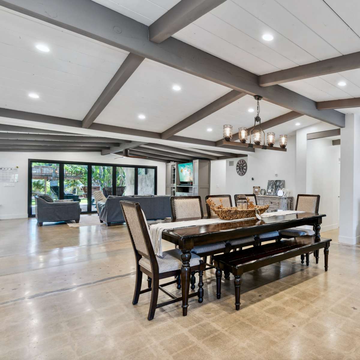 An open-concept dining area featuring a long wooden table with chairs, surrounded by a living space with a seating area, all illuminated by natural light.