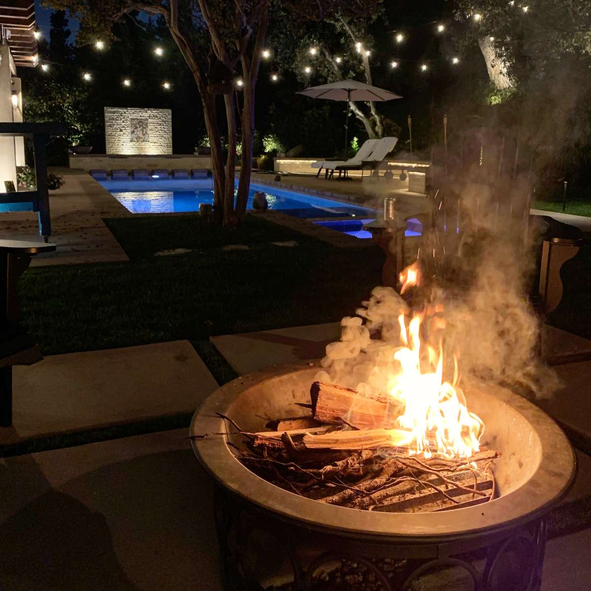 A backyard scene at night features a fire pit with flames, surrounded by stone pathways, a swimming pool, and softly lit trees.