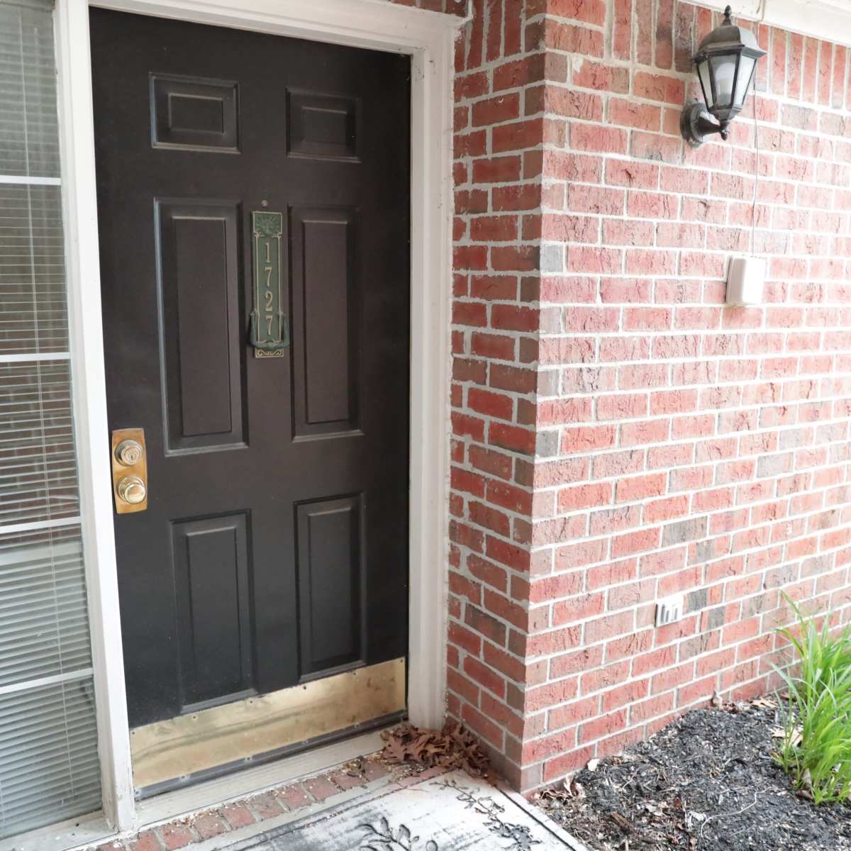 A black front door is framed by brick walls, with a small light fixture and green plants beside a welcome mat.