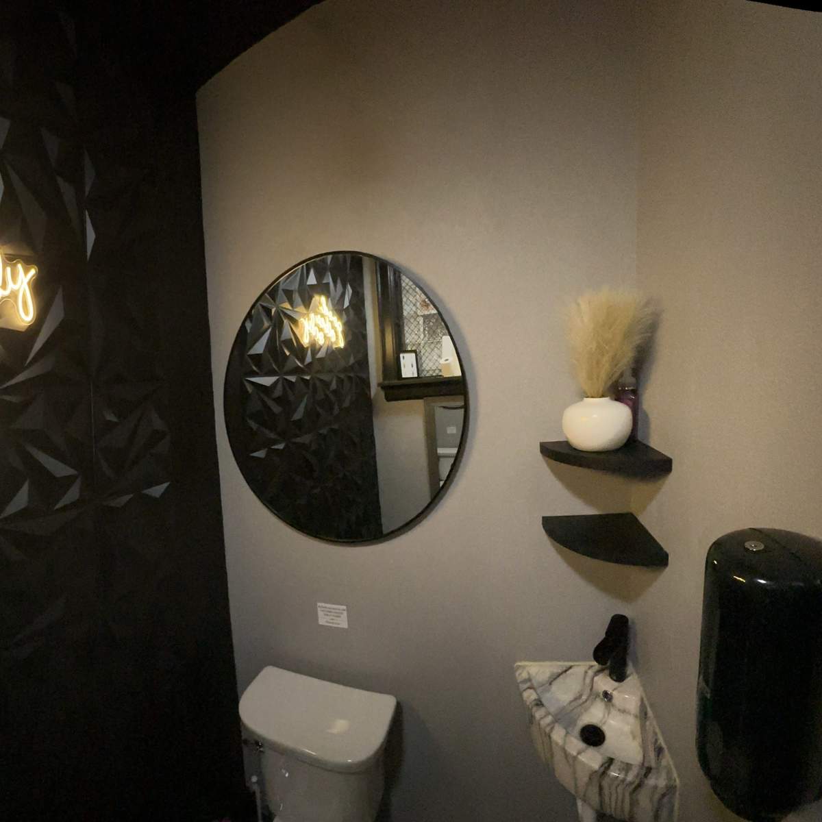 A modern bathroom featuring a black geometric wall, a round mirror, a white toilet, and a neon sign that reads "good vibes only."