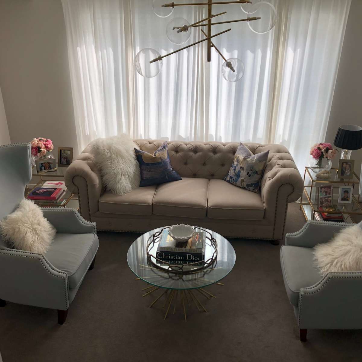 A cozy living room with a beige tufted sofa, two light blue chairs, a glass coffee table, and bright curtains.