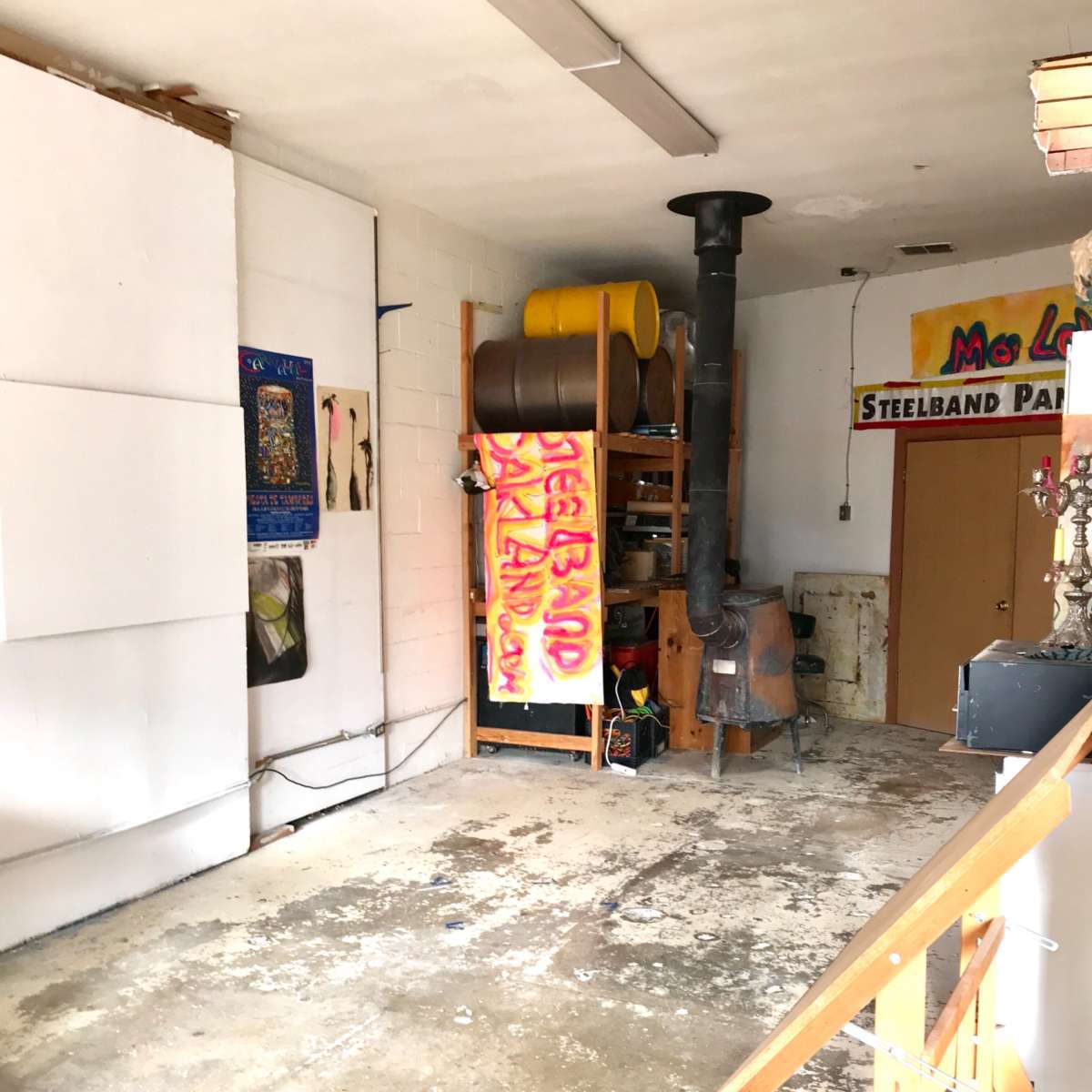 A partially empty garage or workshop with a concrete floor, shelving units, and a wood stove in the corner, along with colorful artwork hung on the walls.