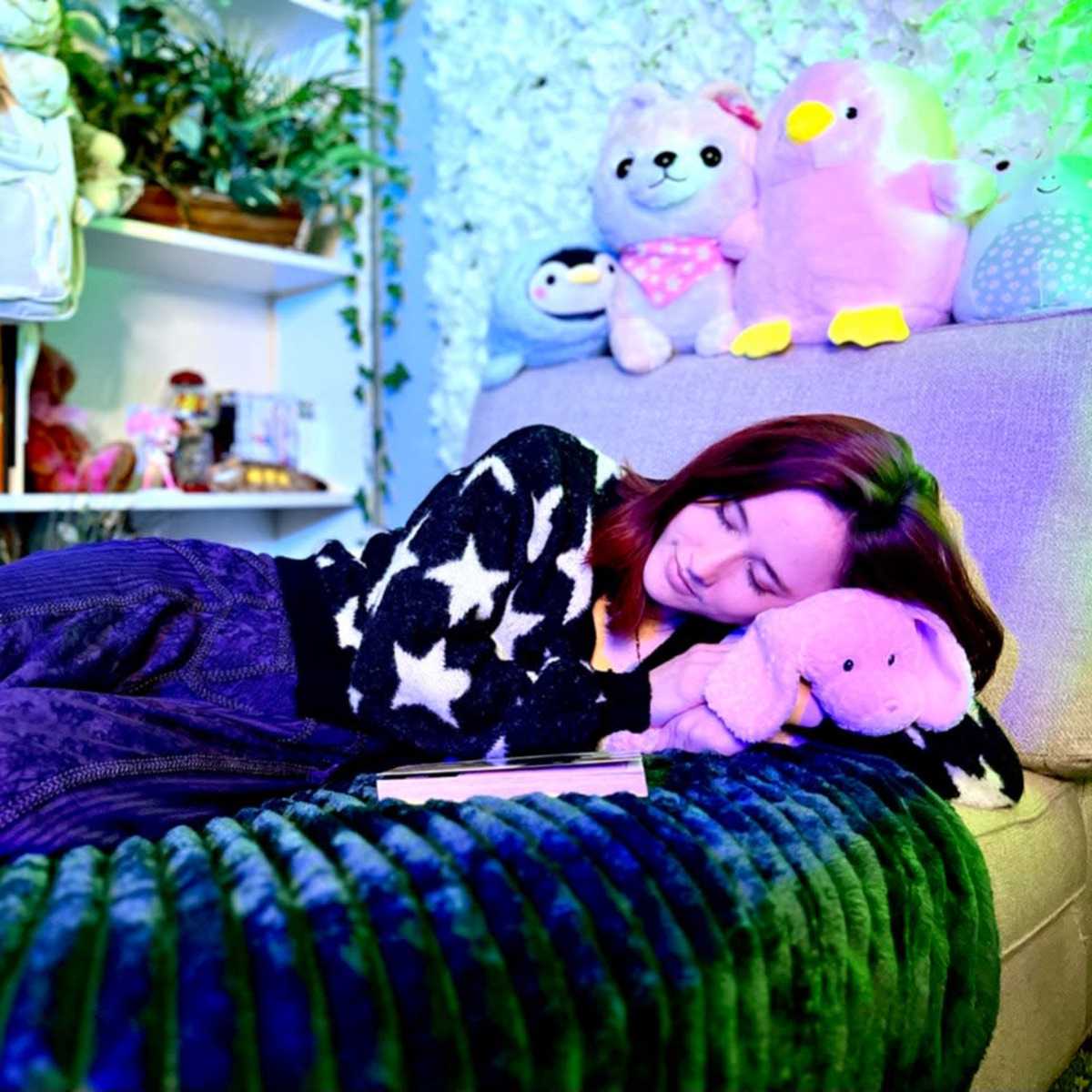 A person is lying on a couch surrounded by plush toys and a bookshelf in a brightly colored room.