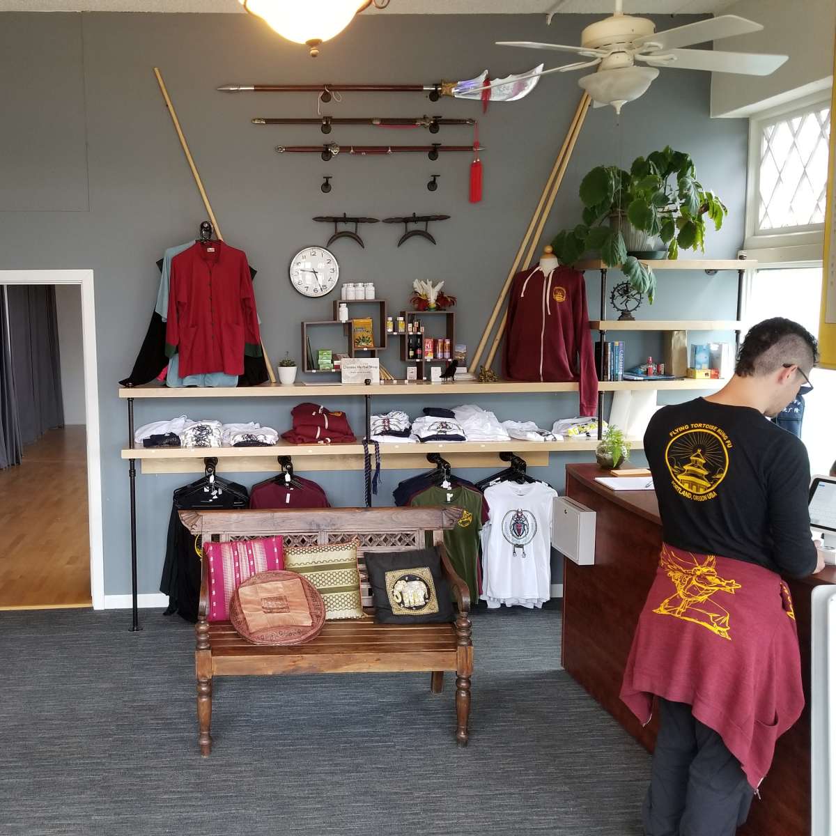 The interior of a retail shop featuring clothing displays on a wooden rack, wall-mounted swords, and a counter where a person is working.