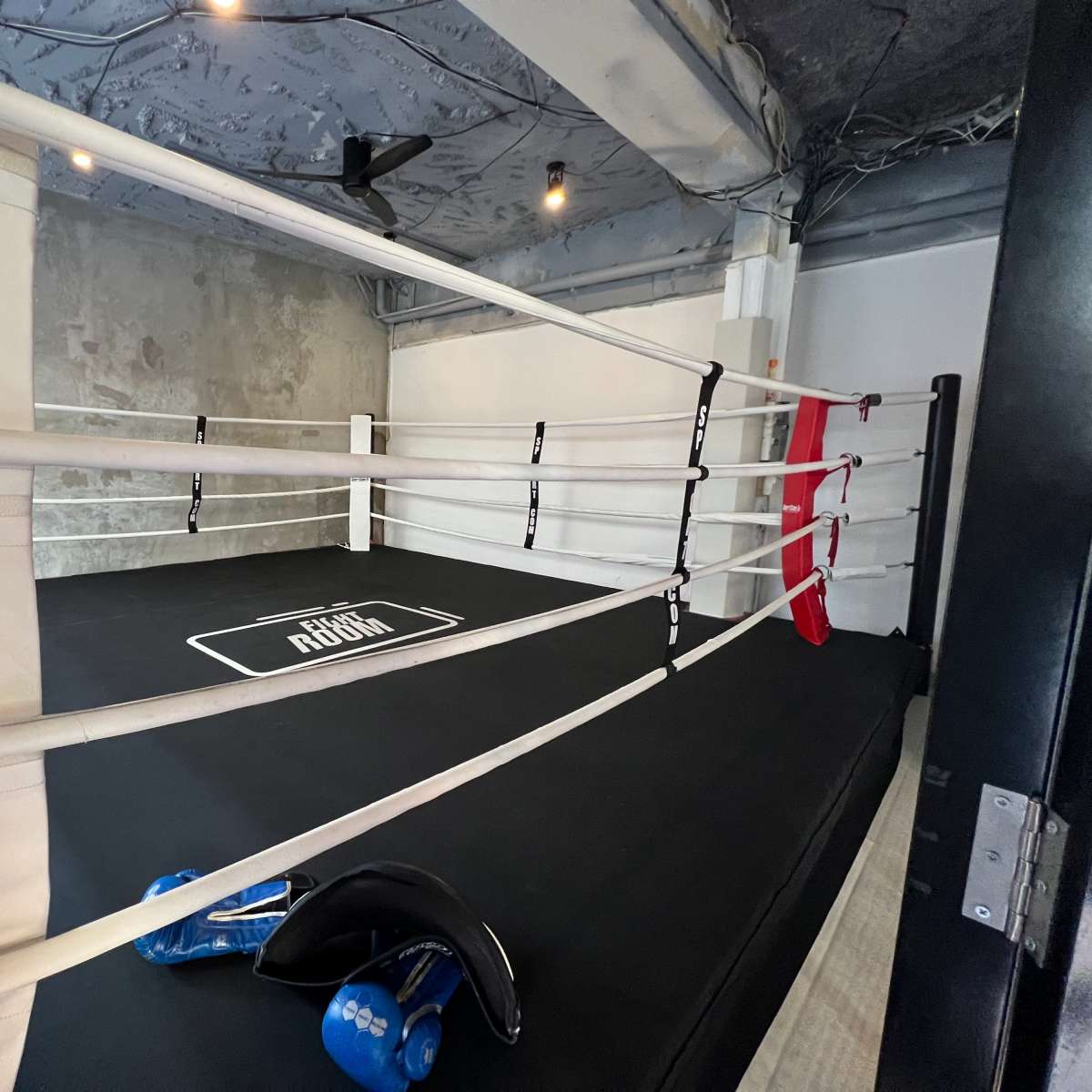 A boxing ring with white ropes and a black mat in a gym setting, accompanied by training equipment on the floor.