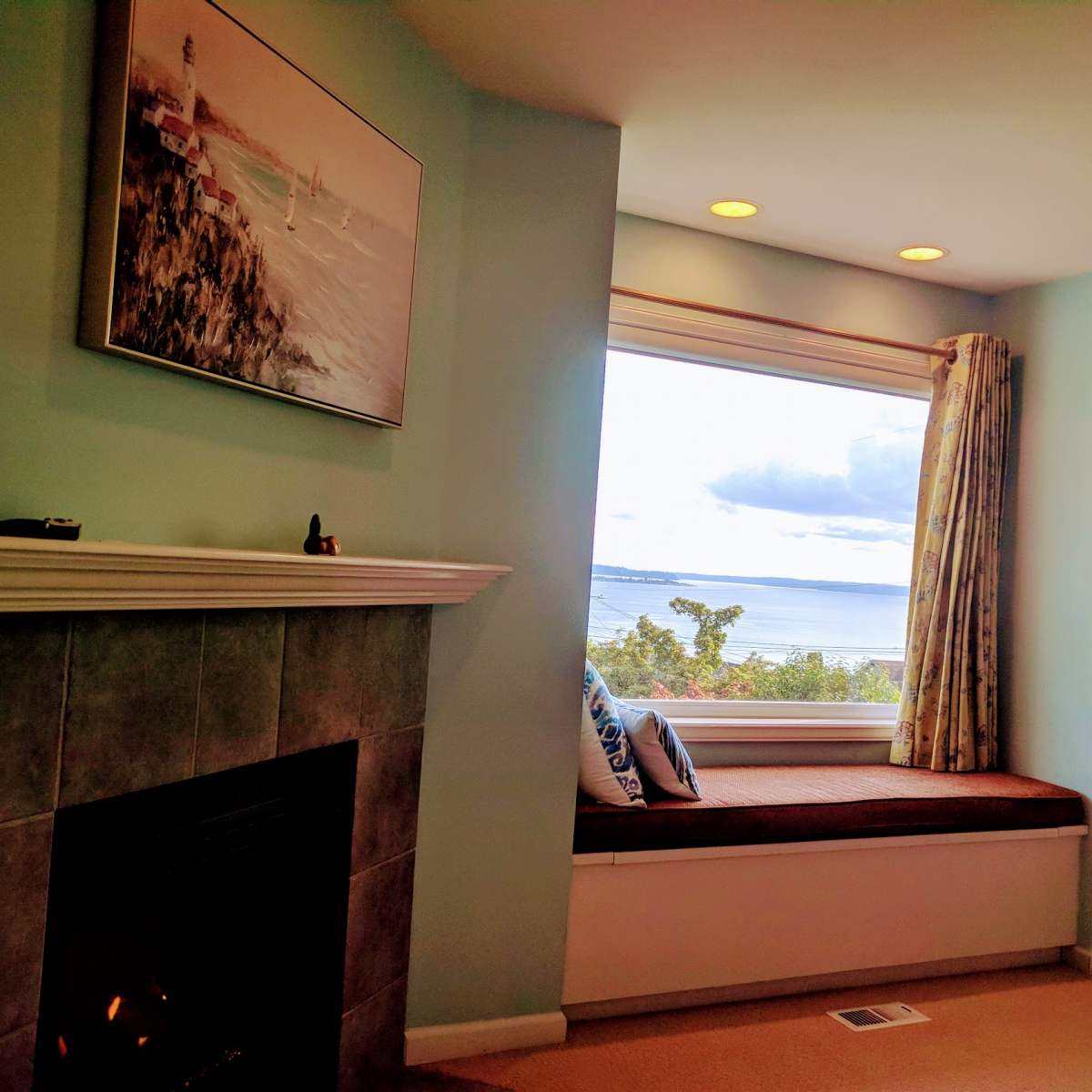 A bright room with a decorative fireplace, a comfortable window seat, and large windows offering a view of the water and sky.